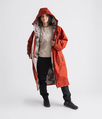 A woman modeling a long hooded red waterproof Changing robe, with a gray and white patterned lining, worn over a beige sweatshirt, black pants, and black boots, against a light gray studio background.