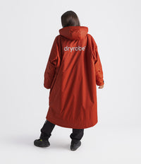 A person shown from the back wearing a long hooded orange-red dryrobe Changing robe, with a white 'dryrobe' logo on the back, paired with black pants and black shoes, against a light gray studio background.
