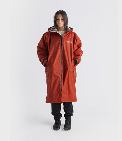 A woman modeling a rust-orange hooded Changing robe, with a front zipper, black cuffs, and a white logo on the chest, standing against a light gray studio background.