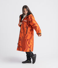 A woman modeling a bright orange hooded Dryrobe Changing Robe with a dark abstract camouflage-like pattern, black zippers, and the brand name 'cryoblue' printed on the chest, set against a plain light gray studio background.