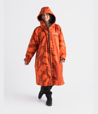 A woman wearing a bright orange hooded Dryrobe Changing Robe with a dark abstract camouflage pattern and the brand name 'dryvoda' on the chest, standing against a light gray studio background.