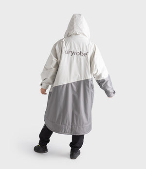 A person shown from the back wearing a hooded Dryrobe in off-white and gray, with black pants and shoes, against a light gray studio background