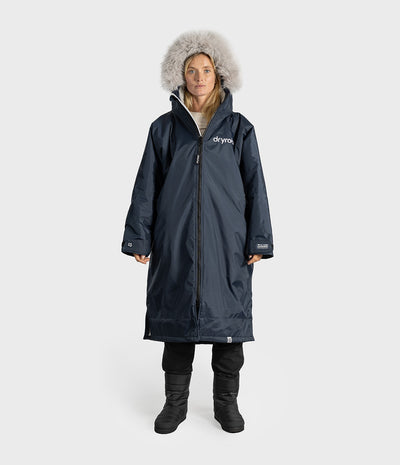 A woman modeling a long navy winter Dryrobe with a large faux-fur trimmed hood, front zipper, and white Dryobe logo on the chest, photographed against a light gray studio background.