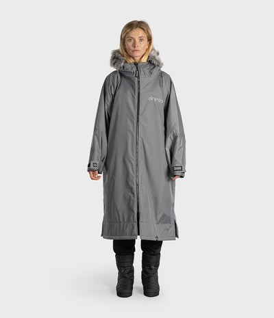 A woman modeling a long gray hooded dryrobe with faux-fur trim, front zipper, and a visible Dryrobe logo on the chest, photographed against a light gray studio background.
