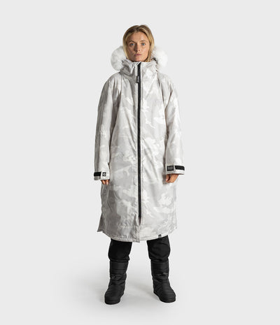 A woman modeling a long hooded winter Dryrobe with a light gray and white camouflage pattern, black zipper, and a fluffy white fur-trimmed hood, paired with black pants and boots against a light gray studio background.