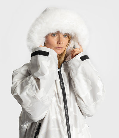 A woman wearing a white camouflage winter Dryrobe with a fluffy white faux-fur hood, photographed against a light gray studio background.