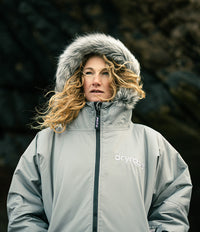 A woman wearing a light gray Dryrobe hooded jacket with a faux-fur trimmed hood, standing outdoors against a dark, blurred natural background.