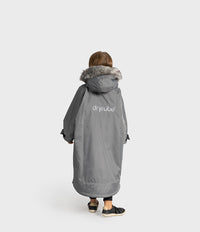 A child viewed from behind wearing a long gray hooded Dryrobe with a faux-fur trim and the brand name 'dryrobe' printed on the back, against a light gray studio background.