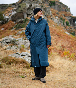 A woman wearing a long navy blue waterproof coat, black beanie, and black trousers stands outdoors in a rocky, grassy mountain setting. The brand name 'Dryrobe' is visible in light blue text on the left chest of the coat.