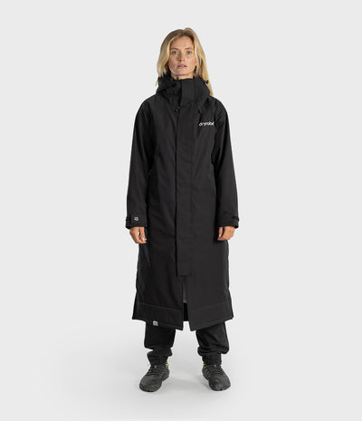A woman modeling a long black hooded outdoor jacket with a high collar and a small white Arktis logo on the left chest, standing against a light gray studio background.