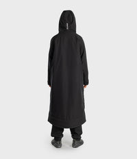 A person shown from the back wearing a long black hooded coat or robe with black pants and dark shoes, standing against a plain light gray studio background.