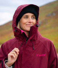 A woman wearing a maroon Dryrobe hooded rain jacket and a black knit beanie, standing outdoors against a soft-focus grassy hillside background.