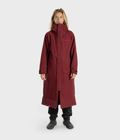A woman modeling a long, dark red hooded technical jacket with black pants and boots, standing against a light gray studio background.