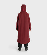 A person shown from the back wearing a long hooded coat in a deep burgundy or maroon color, paired with black pants and dark shoes, against a plain light gray studio background.