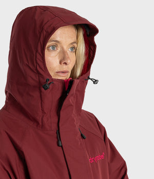 A close-up of a woman wearing a hooded technical jacket in a deep burgundy color, with a small pink 'drymile' logo on the left chest, against a light gray background.