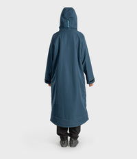 A person shown from the back wearing a long hooded waterproof coat in a muted blue-grey color, paired with black pants and dark shoes, against a plain light grey studio background.