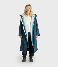 A woman modeling a long navy hooded parka with a white sherpa-style lining and bright turquoise zipper accents, worn open over a cream sweater, black pants, and dark boots against a light gray studio background.