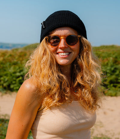 A smiling woman outdoors wearing a black knit beanie, round tortoiseshell sunglasses, and a beige ribbed tank top, with dunes and greenery blurred in the background.