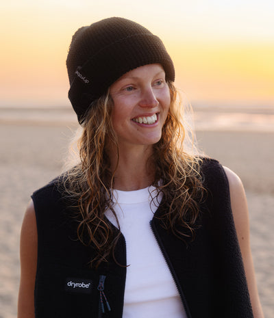 smiling woman wearing a black beanie with 'dryrobe' logo and sunset in the background 