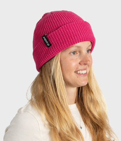 A woman with long blonde hair wearing a bright pink ribbed knit beanie hat. The beanie has a small black fabric logo patch on the side with the brand name 'dryrobe' visible.