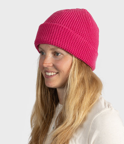 A woman with long blonde hair wearing a bright pink ribbed knit beanie hat, shown in profile against a light gray studio background.