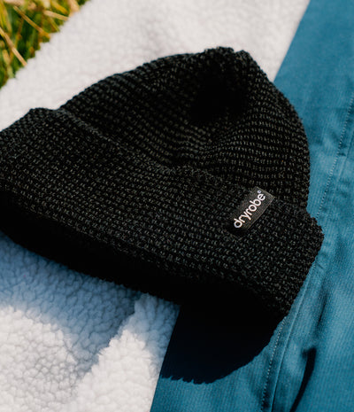 A black ribbed knit beanie with a small black brand tab reading 'dryrobe' in white text, styled on a white sherpa fabric next to a teal garment.