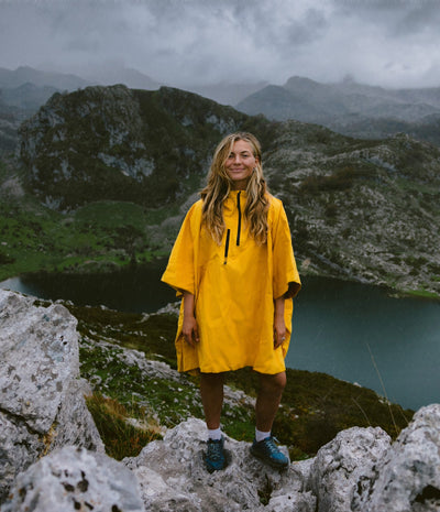 A woman stands on rocky mountain ground with a lake and misty mountains in the background, wearing a bright yellow rain poncho and blue hiking shoes.