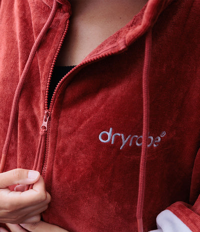 Close-up of a person wearing a rust red fleece zip-up hoodie with white embroidered 'dryrobe' branding on the chest, showing the textured fabric and zipper.