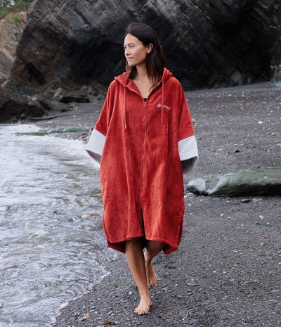 A woman walking barefoot on a rocky beach wearing a terracotta red hooded towel poncho with white sleeve accents. The brand name 'outdoor' is printed in white on the left chest.