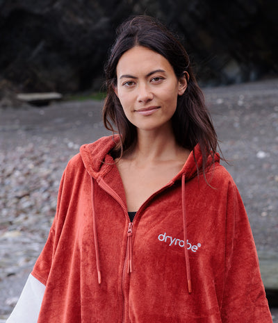 A woman wearing a rust-orange fleece zip-up hooded towel by Dryrobe, standing outdoors on a rocky beach or stream. The white 'dryrobe' logo is printed on the left chest.