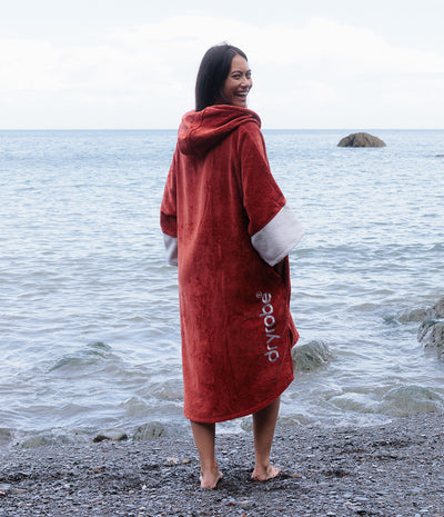 A woman standing on a pebble beach facing the ocean, wearing a red hooded dry robe with white cuffs and a white vertical 'dryrobe' logo.
