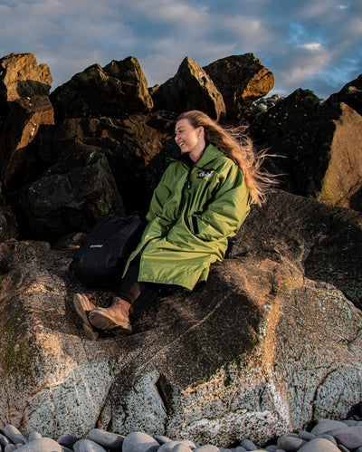 Woman sat on rocks on a beach, wearing dryrobe Advance Long Sleeve REMIX Range change robe in Forest Green Black Camo