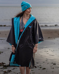 Woman stood in front of the sea in swimming costume, wearing Black Blue dryrobe® Advance Short Sleeve