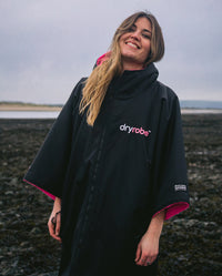 Woman stood smiling on a beach, wearing Black Pink dryrobe® Advance Short Sleeve zipped up 
