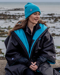 Woman sat on beach wearing Black Blue dryrobe® Adavnce  and Blue dryrobe® Eco beanie  