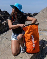 Woman sat in sun on rock, reaching into dryrobe Compression Travel Bag in Orange