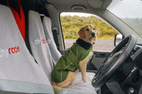 A golden retriever dog sits in the driver's seat of a vehicle wearing an olive green dog coat with a black neck warmer. The car interior has white and gray protective seat covers with the brand name 'drift' partially visible.