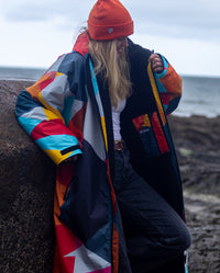 Woman leaning on boulder on a beach, wearing dryrobe Advance Abstract and Beanie 