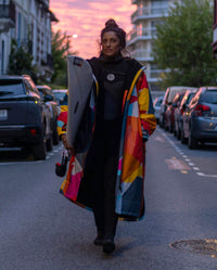 Woman stood in a street in front of sunset, wearing wetsuit and dryrobe Advance Abstract, while carrying surfboard