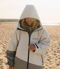 A woman wearing a Dryra hooded Dryrobe stands on a sandy beach at sunset. The jacket is off-white on the upper body with gray lower panels and black zipper details.