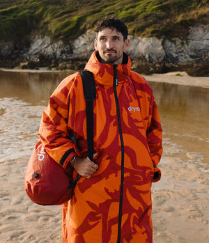 *MALE* A man standing on a sandy beach wearing a bright orange and red patterned Dryrobe Changing Robe, with a red Dryra bag slung over his shoulder, against a rocky shoreline background.