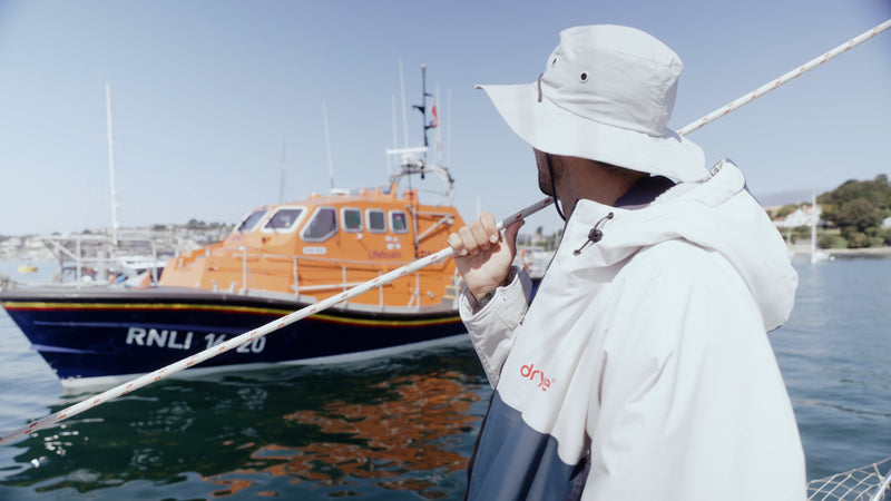 A person wearing a white hooded dryrobe and a white bucket hat stands on a boat in a harbour, holding a long pole. An orange and blue RNLI life boat is visible in the background under a clear blue sky.