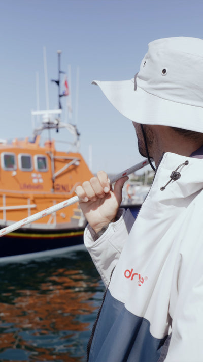A man wearing a white sun hat and a white and navy Dryrobe with a red 'dryrobe' logo, standing on a dock with an orange boat in the background.