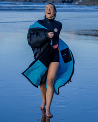 Woman running across a beach, wearing a wetsuit and Black Blue dryrobe® Advance Long Sleeve