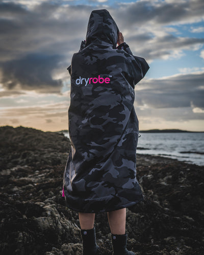 Woman stood on beach facing out to sea, wearing Black Camo Pink dryrobe® Advance Long Sleeve with hood up