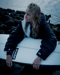 Woman crouching on rocks holding surfboard, wearing Black Grey dryrobe® Advance Long Sleeve 