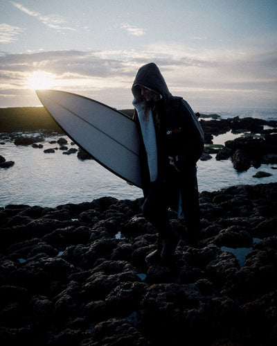 Woman walking across rocks holding surfboard, wearing Black Grey dryrobe® Advance Long Sleeve with hood up