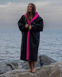 Woman stood on rocks by the sea, wearing Black Pink dryrobe® Advance Long Sleeve