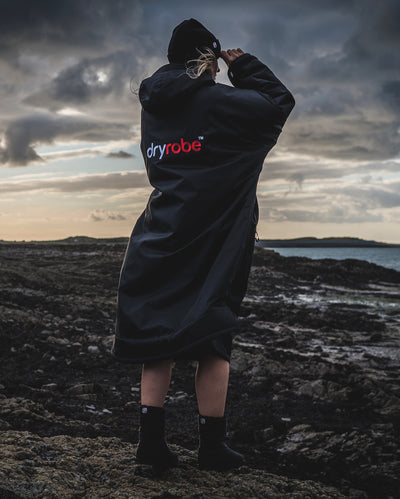 Woman stood on the beach looking out to sea, wearing dryrobe Advance Long Sleeve changing robe in Black Red 