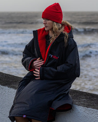 Woman sat on wall in front of sea, wearing dryrobe Advance Long Sleeve changing robe in Black Red and Red dryrobe Beanie